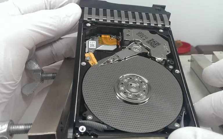 Laboratuvarda eldivenli eller arasında kapağı açılmış hard disk, plaka ve okuma-yazma kafası görünüyor